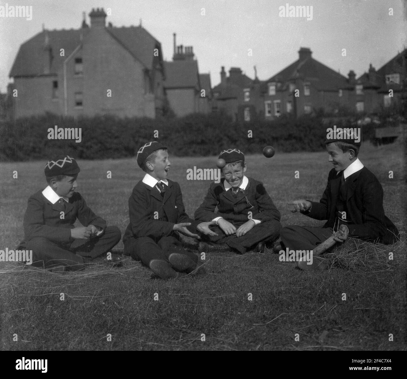 1930s, historisch, sitzen zusammen auf einem Feld draußen, vier englische öffentliche Schuljungen in ihrer traditionellen Uniform und Kappen, spielen "fangen" mit einem Cricket-Ball, England, Großbritannien. Stockfoto