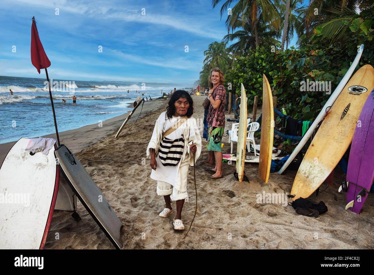 Ein indigener Kogi-Mann überquert mit einem Surfer am Strand in Palomino, Kolumbien, Wege. Stockfoto