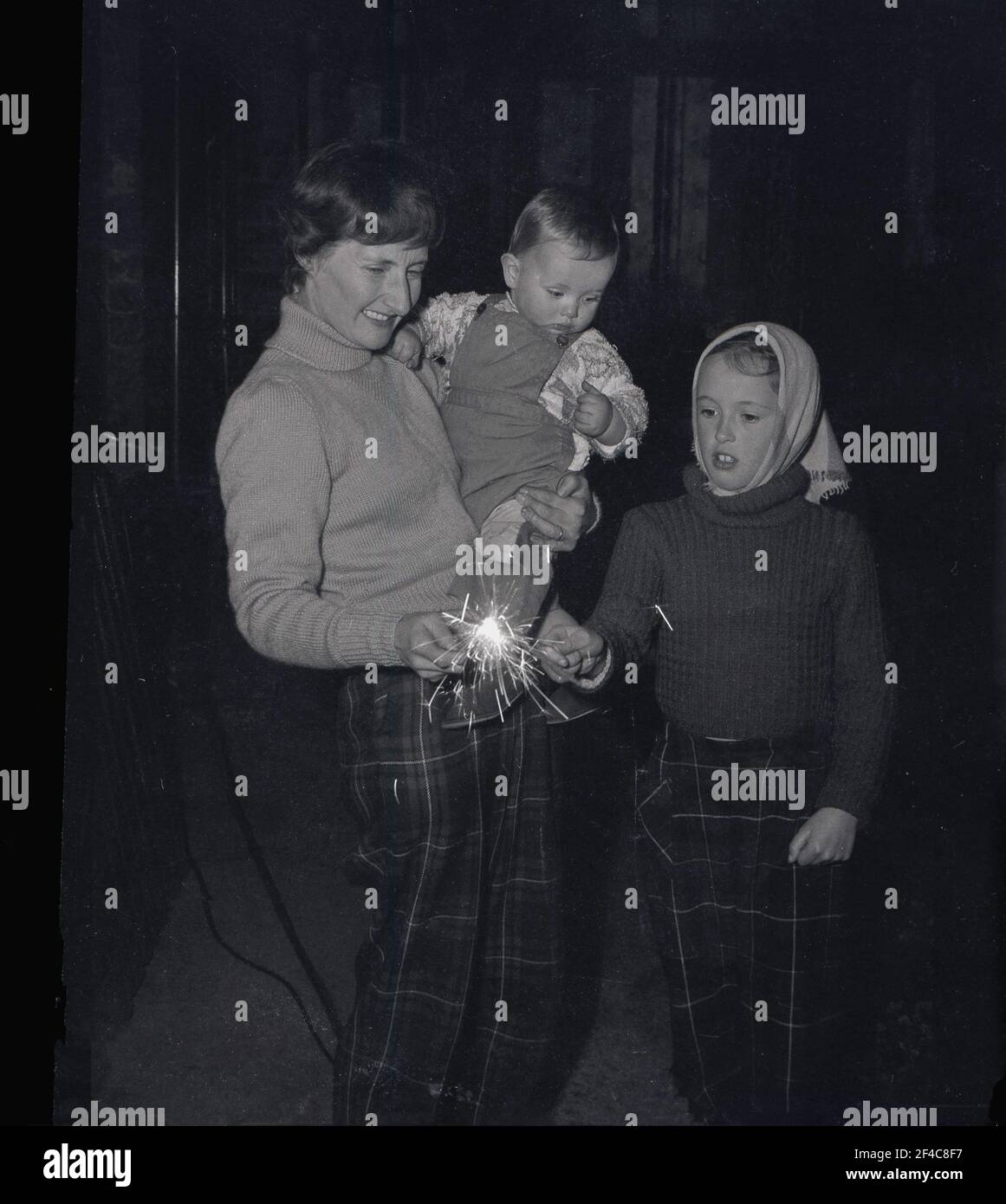 1960s, historische, abendliche und Lagerfeuernacht, und eine Mutter, die ihr Kleinkind in ihrem linken Arm hält und mit Sparkern mit ihrer jüngeren Tochter spielt, die beide eine Tartanhose tragen, Fife, Schottland, Großbritannien. Stockfoto
