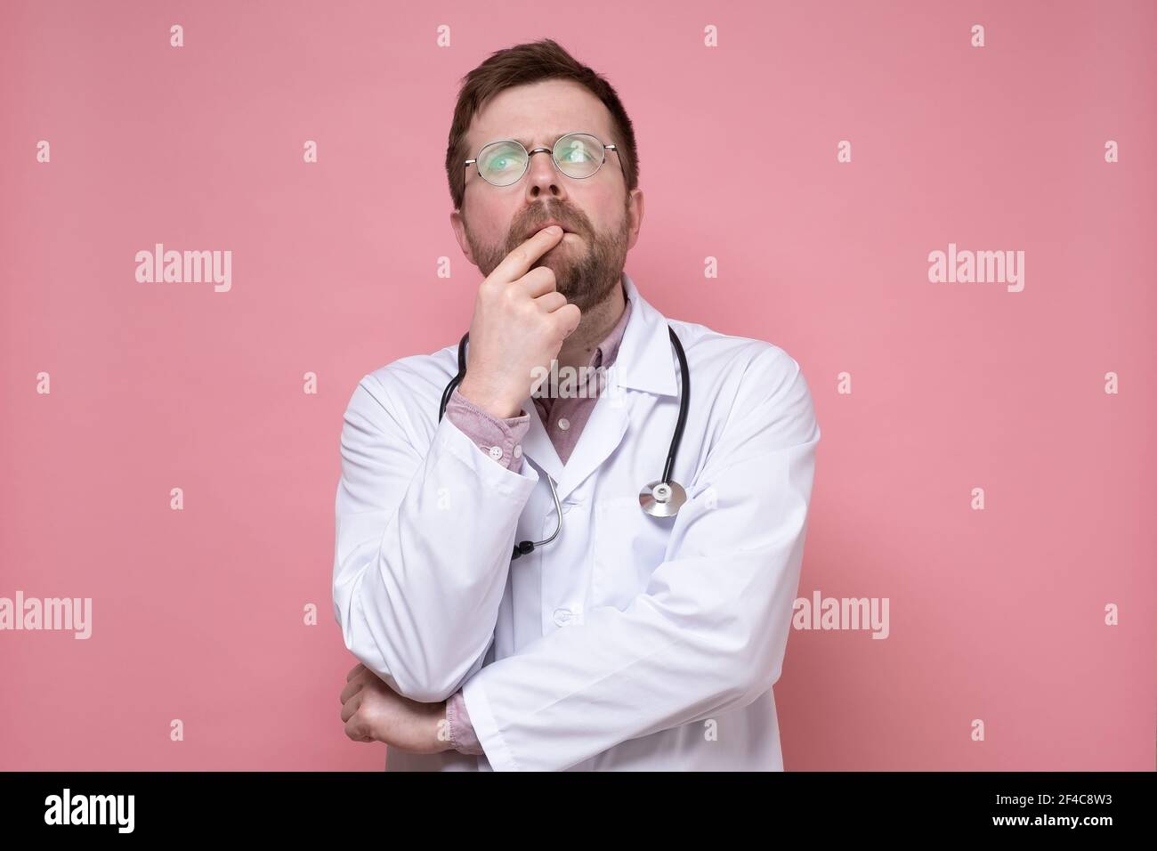 Rätselhafter Arzt mit Brille, einem weißen Mantel und einem Stethoskop um  den Hals sieht nachdenklich nach oben. Rosa Hintergrund Stockfotografie -  Alamy