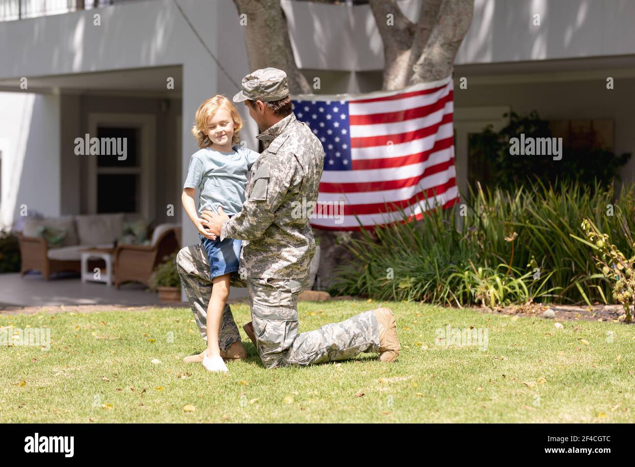 Kaukasischer Soldat Vater knieend umarmenden Sohn im Garten mit amerikaner Flagge hängt vor dem Haus Stockfoto