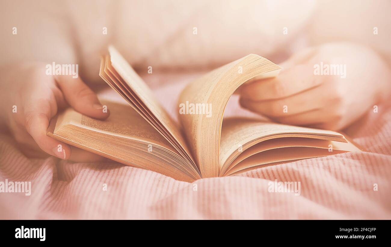 Ein Mann in einem weißen Pullover liegt auf einem Bett mit einer rosa gestreiften Decke und liest ein Interessantes altes Buch im Morgenlicht. Selbstentwicklung und Stockfoto