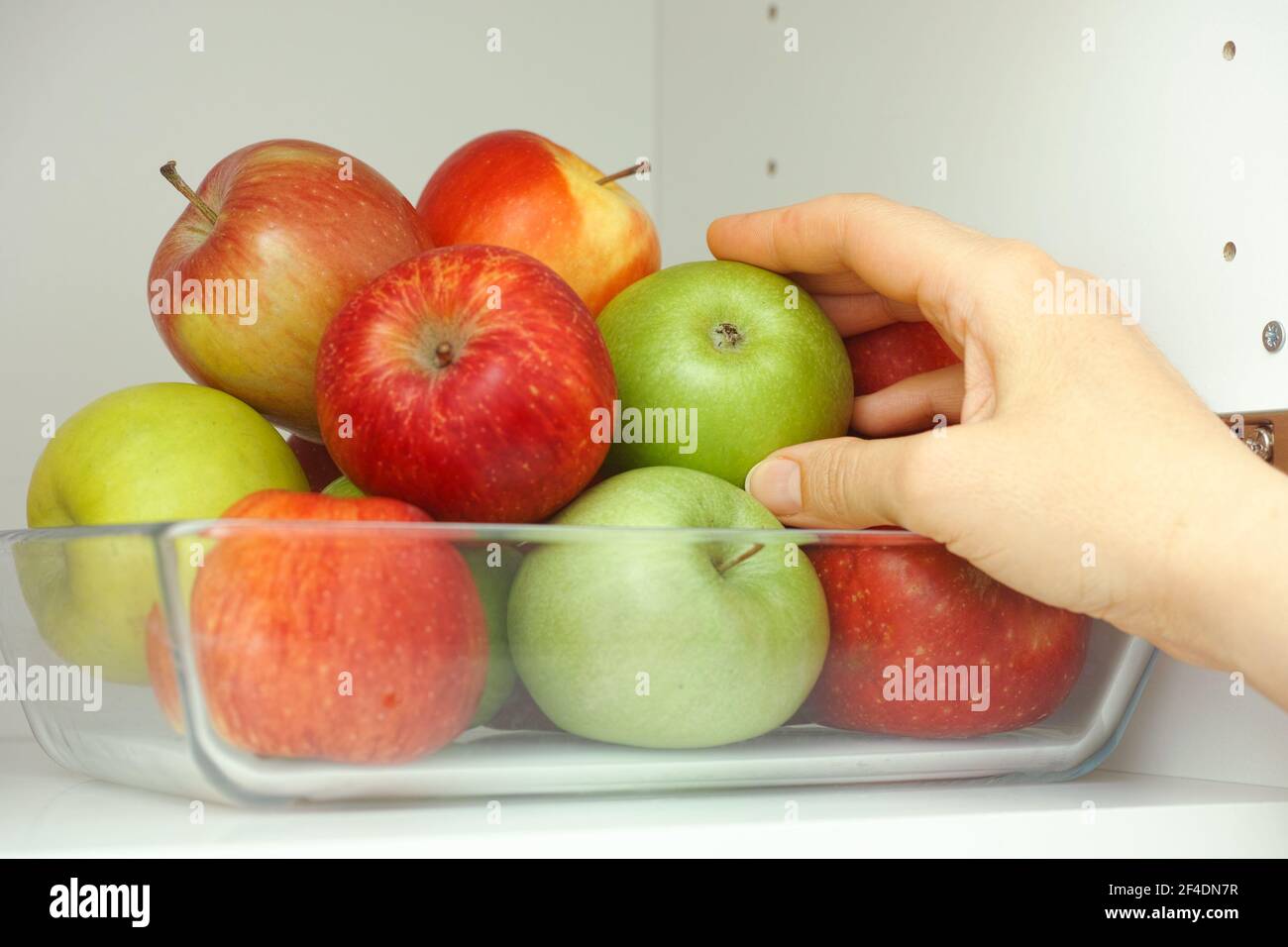 Eine Frau, die einen grünen Apfel aus einem Stapel Äpfel nimmt, die in einem Glasbehälter sind, der in einem Küchenschrank ist. Nahaufnahme. Stockfoto