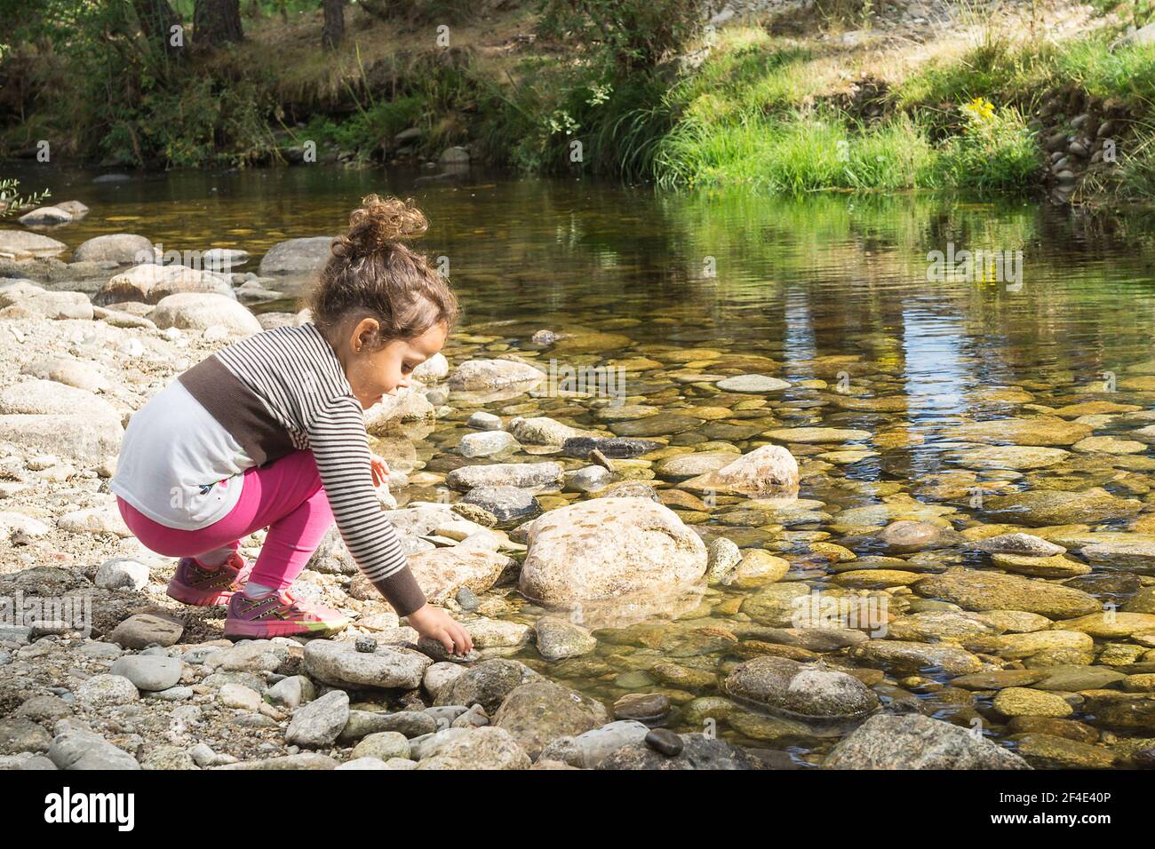 Mädchen mit gesammelten Haaren, rosa Hosen und gestreiften Cardigan im Fluss gebogen über Abholung einen Stein, um es in das transparente Flusswasser zu werfen Stockfoto