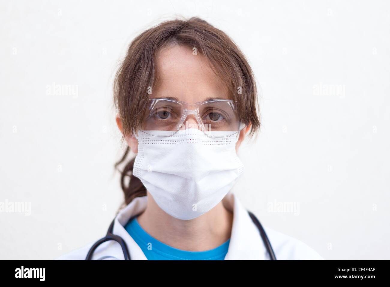 Nahaufnahme der Ärztin mit weißer Maske und Schutzbrille mit gesammeltem Haar, weißem Fell und weißem Stethoskop-Hintergrund Stockfoto