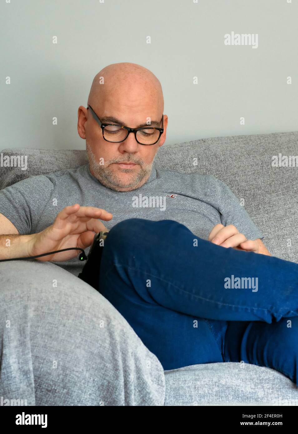 Kahlköpfiger Mann mittleren Alters, der auf dem Sofa sitzt und nach unten schaut Wie er sein iPad benutzt Stockfoto