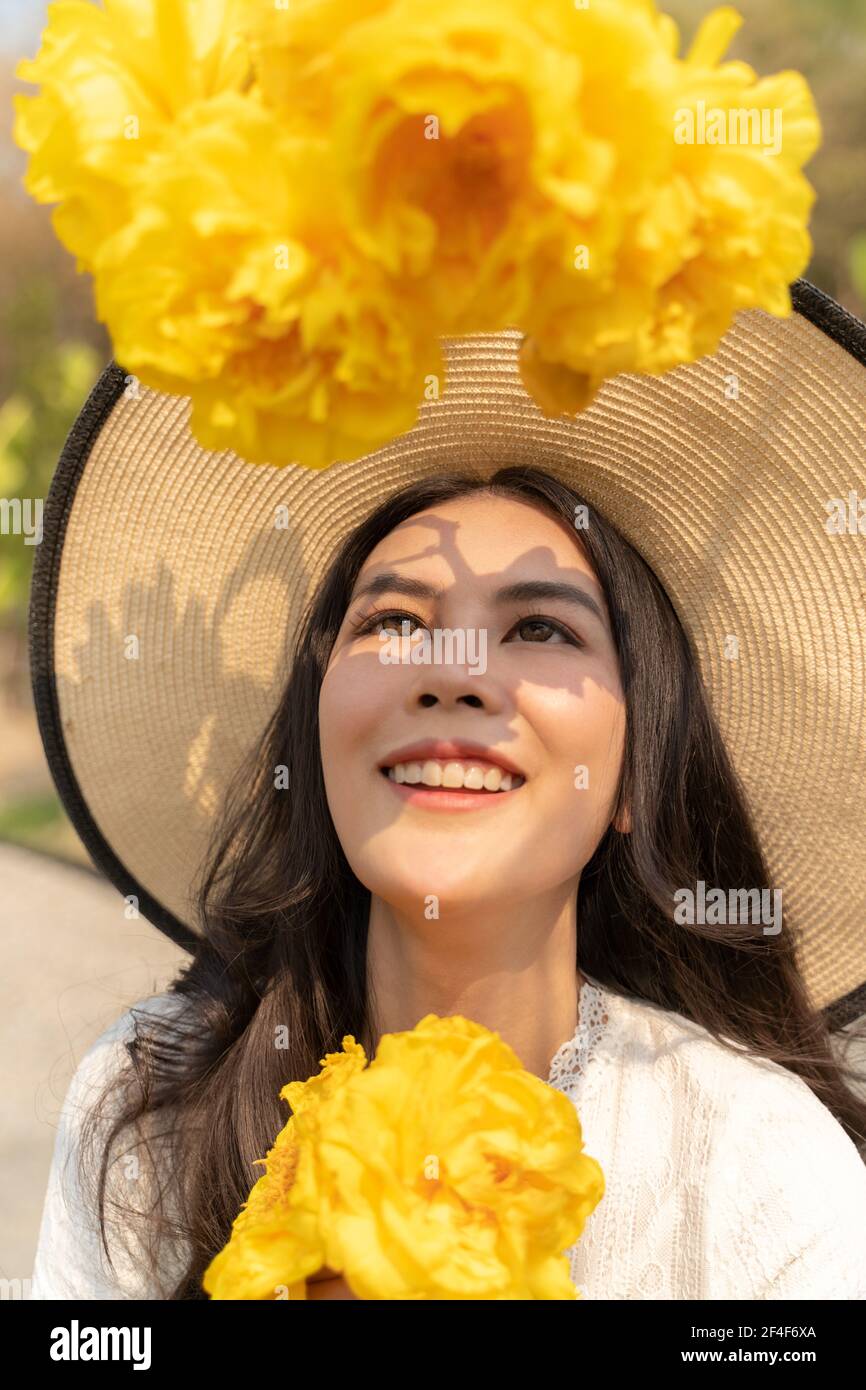 Porträt von glücklichen jungen schönen Frau trägt großen Strohhut und weißes Kleid, lächelnd und Blick auf große gelbe Blume vor ihr auf hellen s Stockfoto