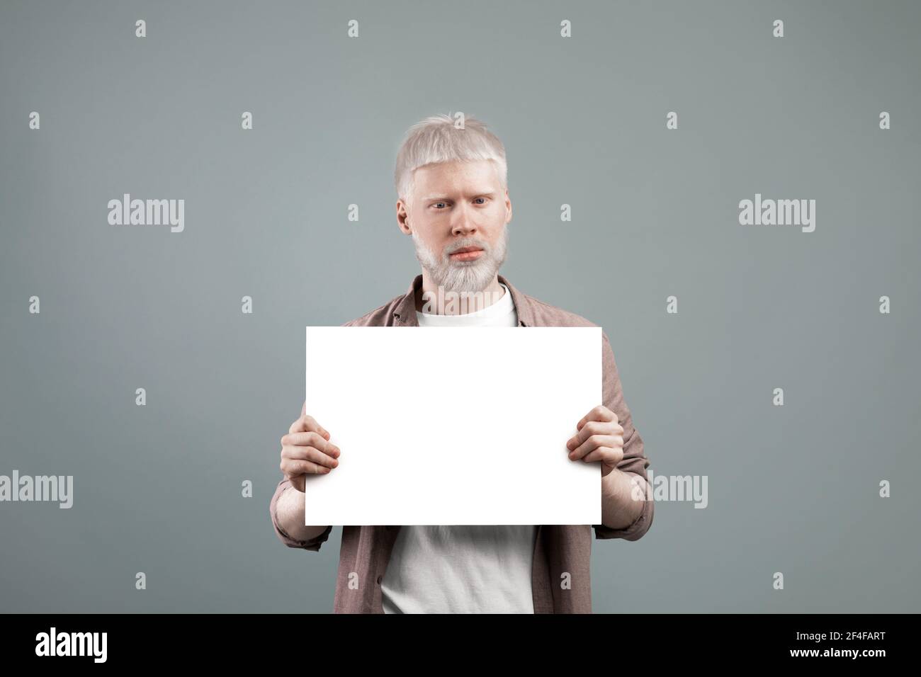 Albino Mann hält leeres Papierblatt mit freiem Platz, posiert vor der Kamera auf grauem Hintergrund, Werbung und Mockup Stockfoto