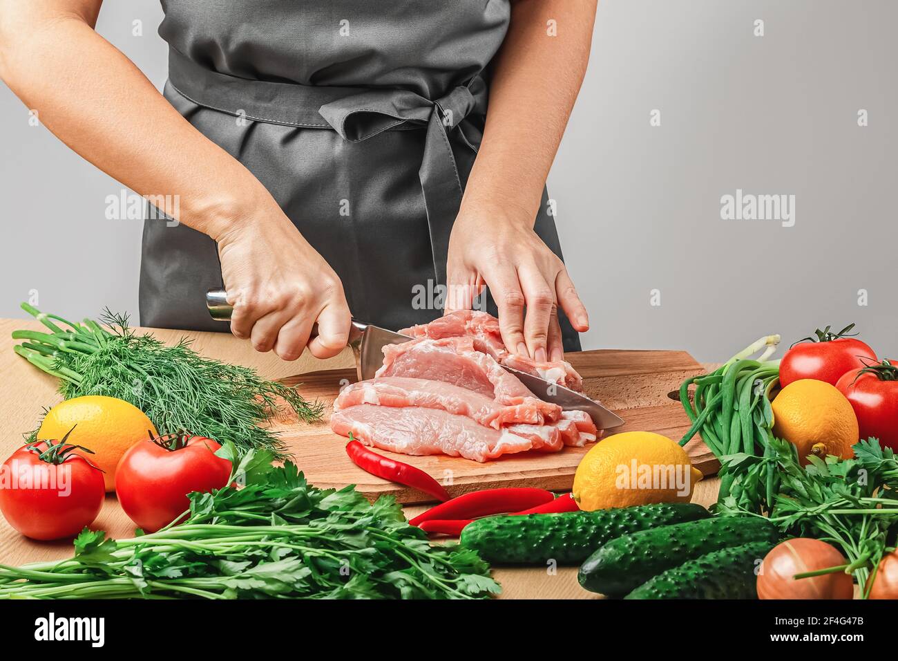 Eine Frau in einer grauen Schürze schneidet ein Stück Fleisch mit einem Messer auf einem Holzbrett. Es gibt Zwiebeln, Tomaten, Dill, Petersilie, Tomaten, Rote Paprika und Stockfoto