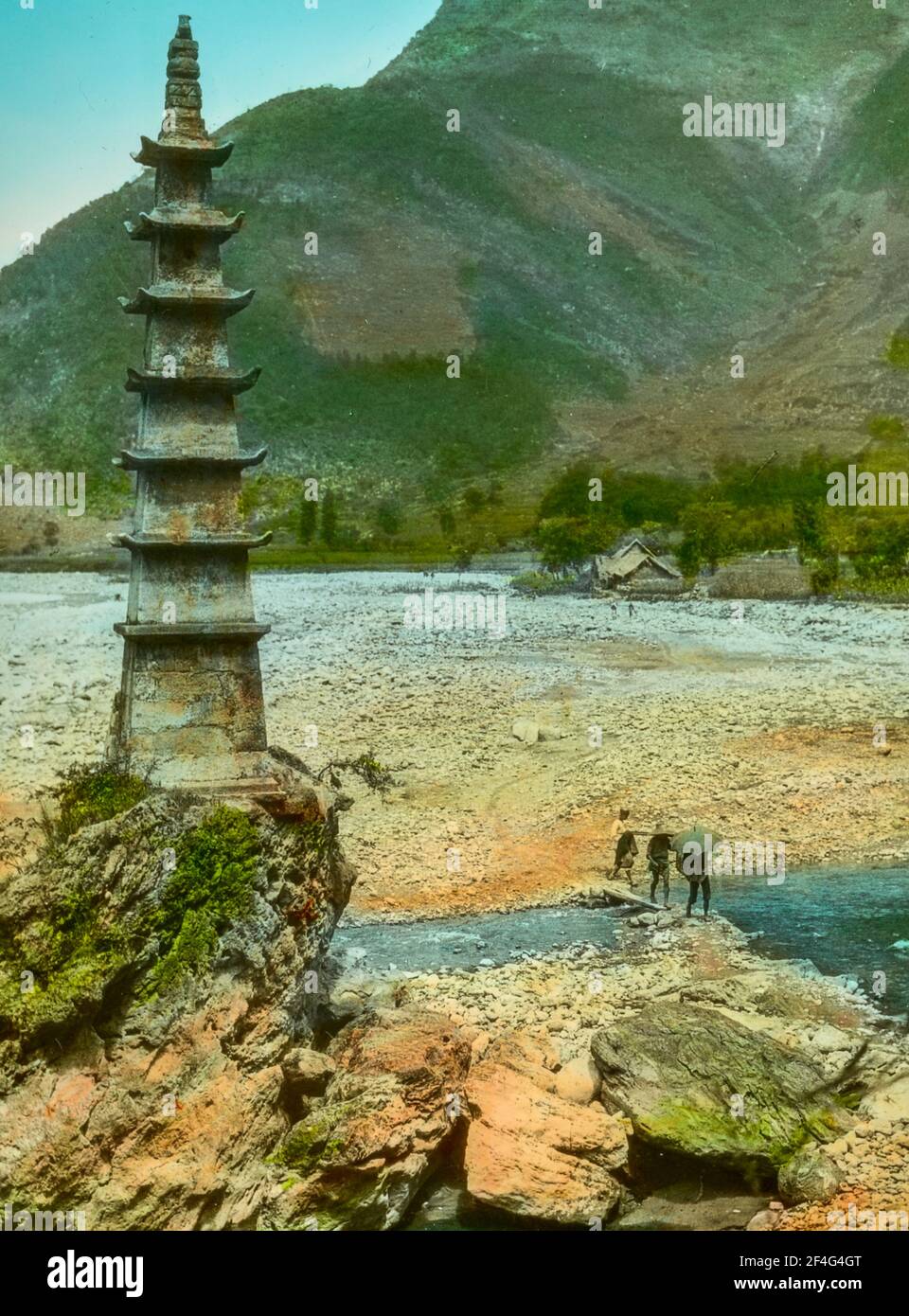 Drei Menschen überqueren an einem sonnigen Tag eine Plankenbrücke über einen teilweise trockenen Fluss, um eine kleine Pagode zu erreichen, die auf einem Felsvorsprung errichtet wurde, Sichuan, China, 1917. Aus der Sammlung Sidney D. Gamble Photographs. () Stockfoto