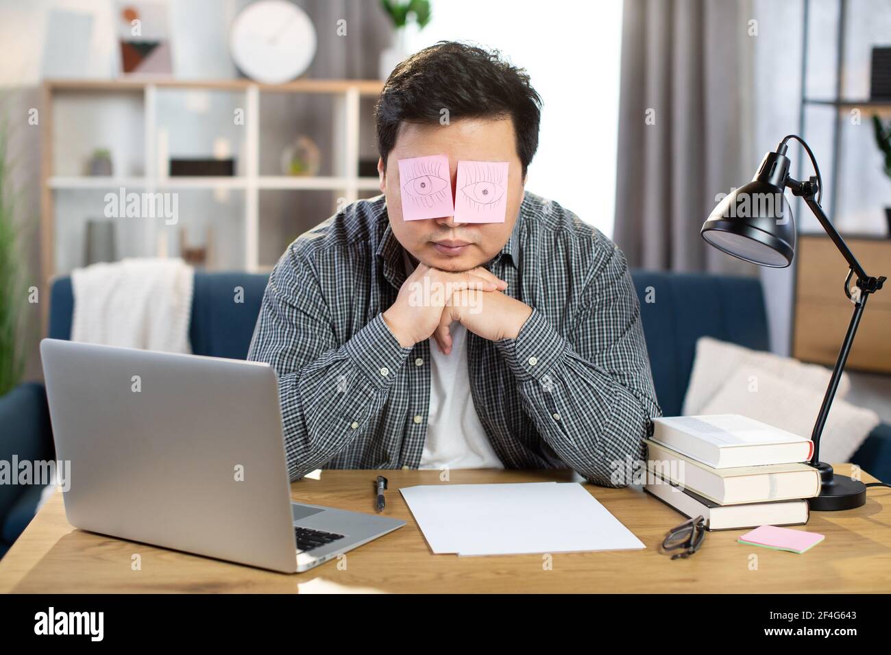 Müde asiatische Geschäftsmann schlafen am Arbeitsplatz mit gefälschten  Augen auf Papier Aufkleber gemalt bedeckt seine Augen. Junger Mann sitzt am  Tisch mit Laptop und lehnt sich an die Hände. Überarbeiteter Begriff  Stockfotografie -