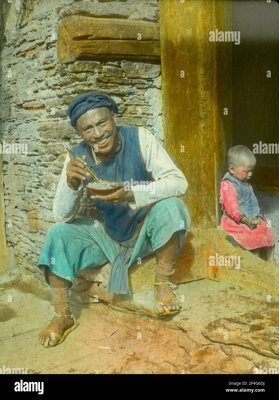 Nahaufnahme eines lächelnden Mannes mit Essstäbchen und einer Schüssel mit Essen, der an einem sonnigen Tag in einer Steintür sitzt, mit einem kleinen Kind im Hintergrund, Sichuan, China, 1917. Aus der Sammlung Sidney D. Gamble Photographs. () Stockfoto