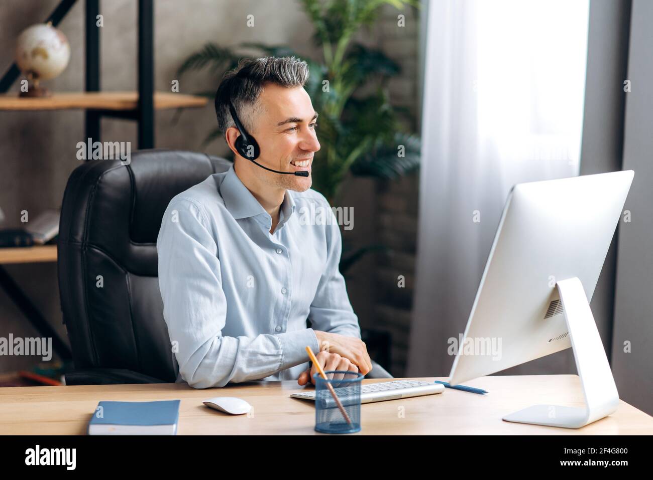 Ein fröhlicher kaukasischer Senior Business Leader, Manager, sitzt an einem Tisch, im Büro, in einem stilvollen Hemd, kommuniziert über einen Videoanruf mit Mitarbeitern oder Freunden, mit einem Headset und Computer, lächelnd Stockfoto