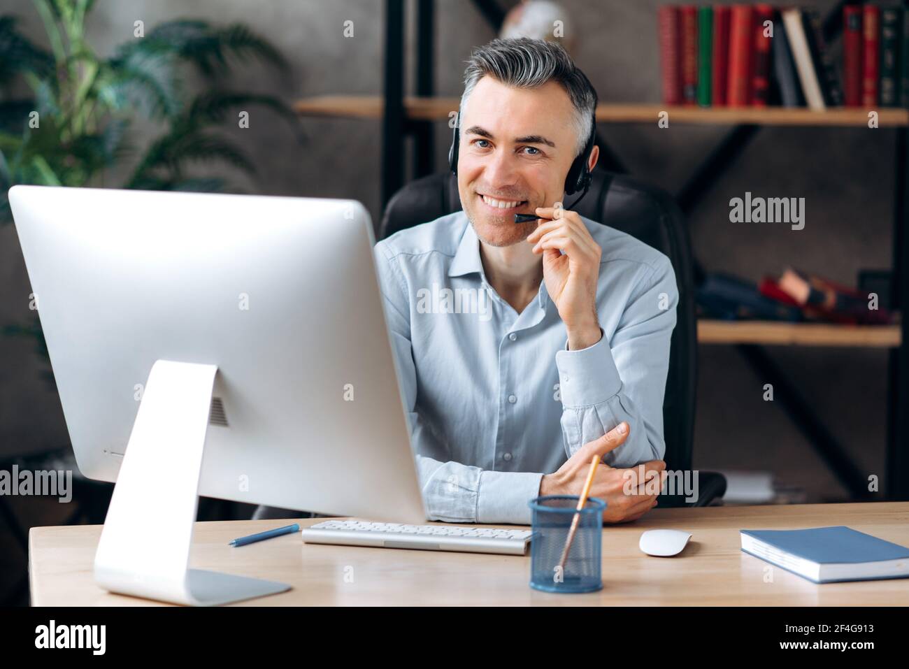 Kaukasische zuversichtlich erfolgreichen Geschäftsmann, Trainer, sitzt an einem Tisch im Büro, trägt Kopfhörer und formelle Hemd, verwendet Computer, arbeitet aus der Ferne , Blick auf die Kamera mit freundlichem Lächeln Stockfoto