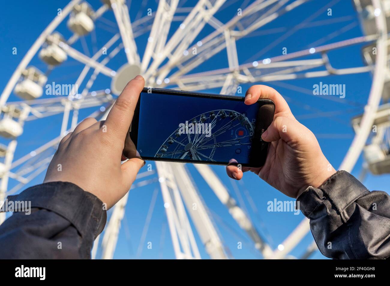 Zamora, Spanien. März 20, 2021. Eine Hand einer Person mit einem Handy macht während des ein Bild von einem weißen Riesenrad mit dem blauen Himmel im Hintergrund Stockfoto