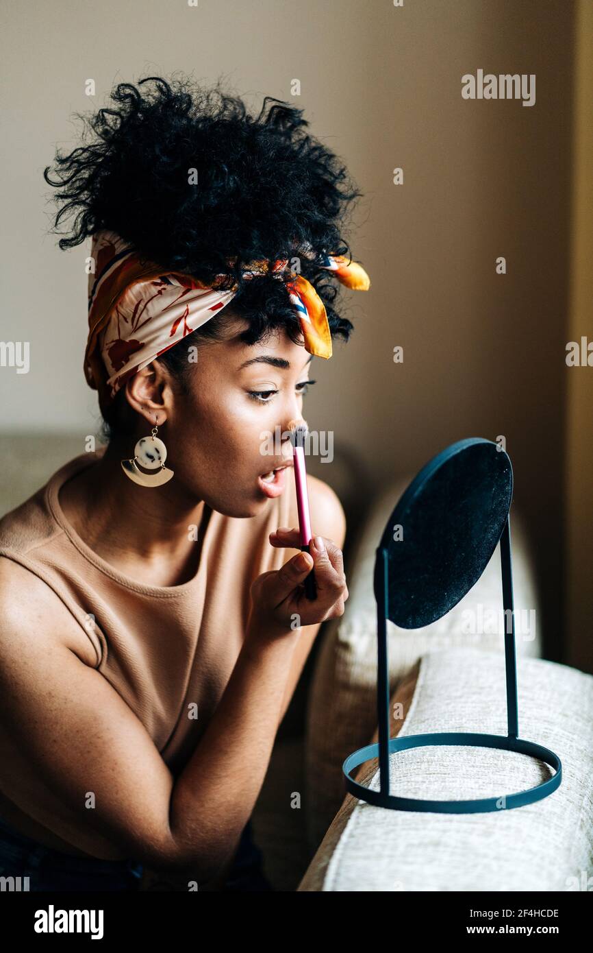 Seitenansicht einer charmanten afroamerikanischen Frau, die in der Runde schaut Spiegel und Auftragen von Pulver auf das Gesicht, während Make-up an Zu Hause Stockfoto