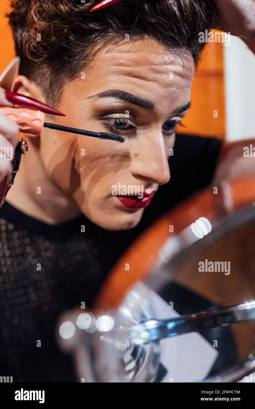Crop junge Queer mit Maniküre berühren Haar beim Auftragen Mascara Auf langen Wimpern gegen Spiegel Stockfoto