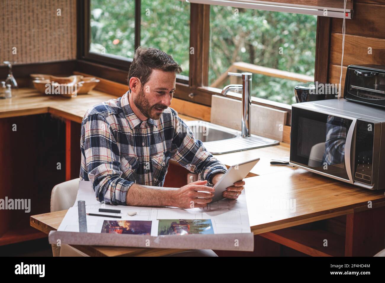 Lächelnder männlicher Architekt mit Blaupause des Hauses, das am Tisch sitzt Zu Hause und mit Videoanruf über Tablet während des Sprechens Über neues Projekt Stockfoto