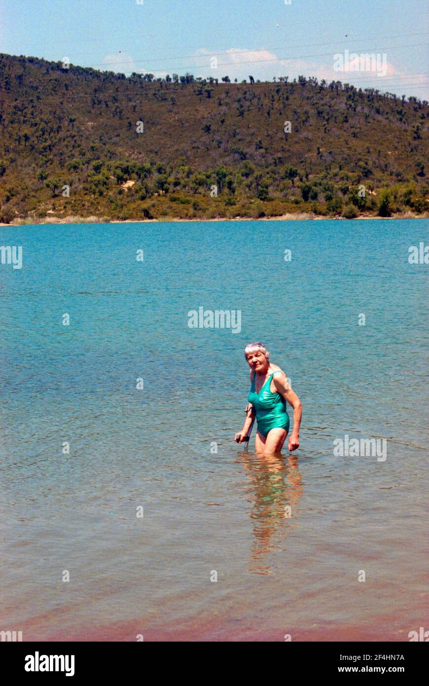Lächelnde ältere Frau im Badeanzug, knietief im türkisfarbenen See stehend, sich auf einer Krücke feststehend. Stockfoto