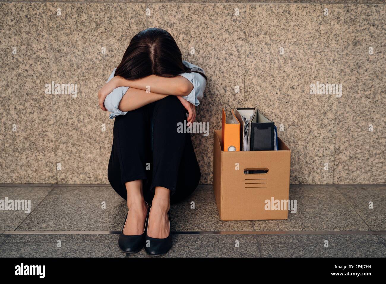 Unbekannte junge Frau, die ihr Gesicht versteckt, nachdem sie entlassen wurde Von der Arbeit wegen der wirtschaftlichen Rezession sitzen mit Karton Habseligkeiten auf dem Boden und weinen Stockfoto
