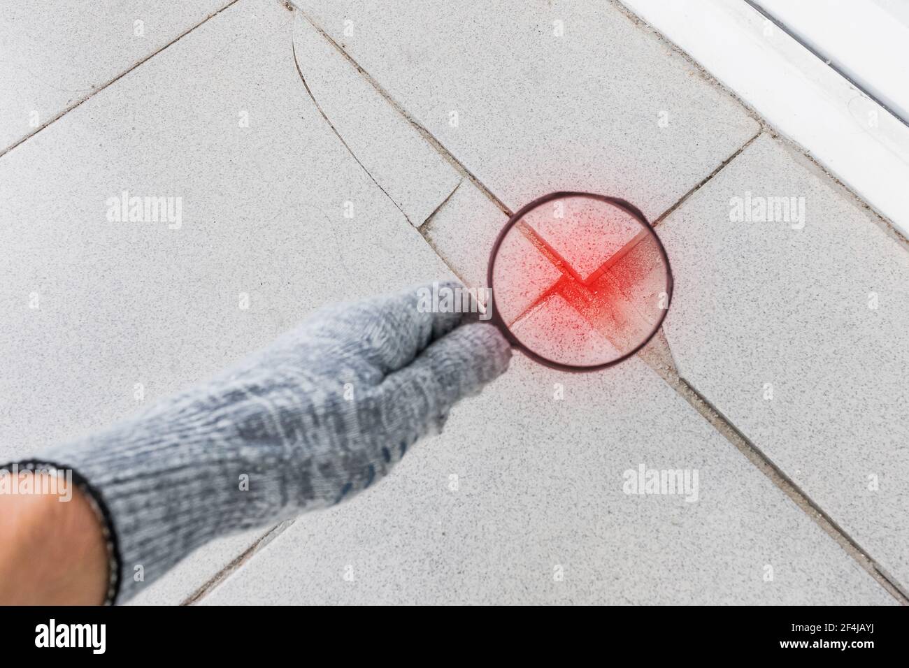 Die Hand eines Industriearbeiters in einem Bauhandschuh mit Lupe untersucht den beschädigten rissigen Fliesenhintergrund. Renovierungskonzept. Stockfoto