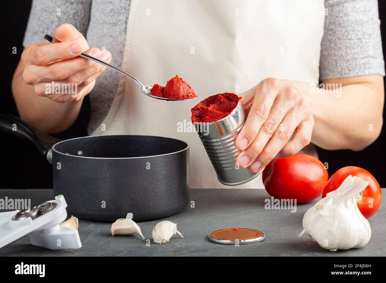 Eine kaukasische Frau fügt Tomatenmark aus einer Dose in ein Gericht hinzu, das sie in einem Antihaft-Topf zubereitet. Es gibt frische Tomaten, Knoblauch, einen Dosenöffner auf Stockfoto