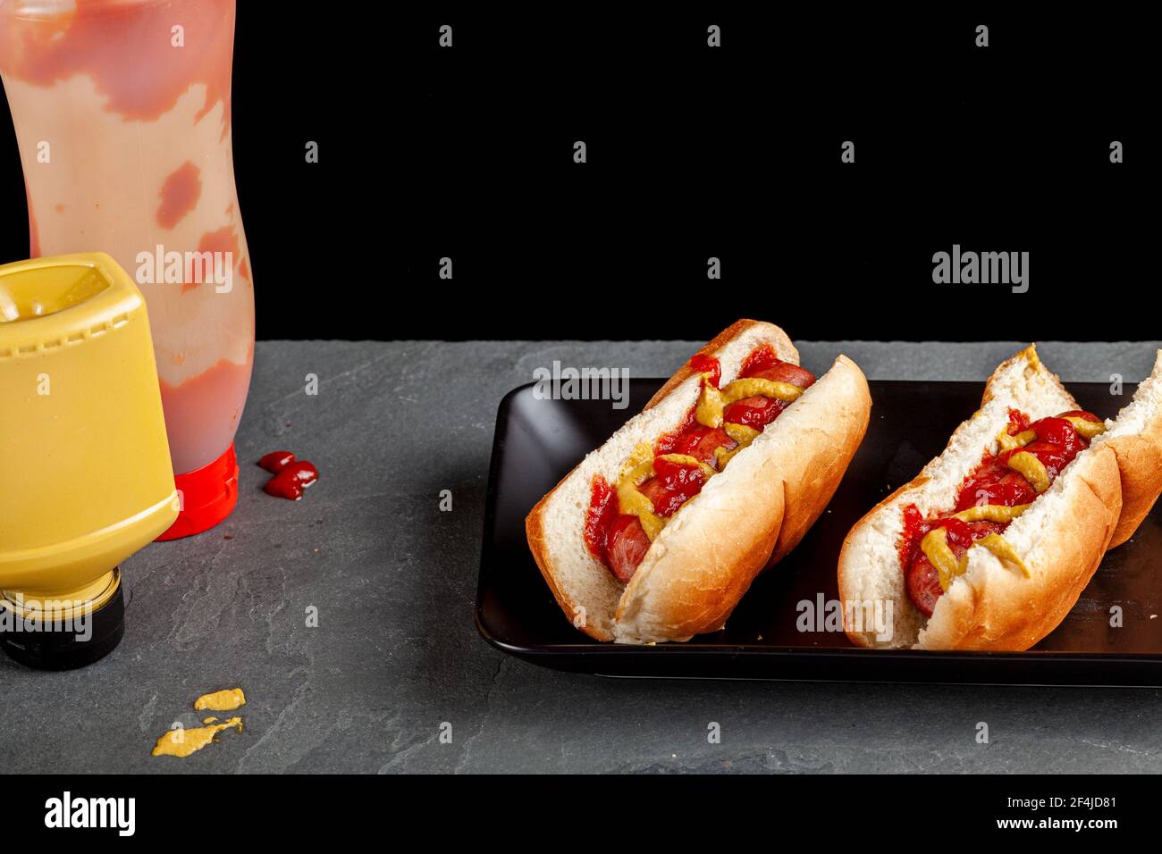 Zwei Hot Dog Sandwiches in speziellen Brötchen und mit Ketchup gekrönt, Senfsoßen werden auf einem flachen schwarzen Teller auf Küchentisch schnell und einfach platziert Stockfoto