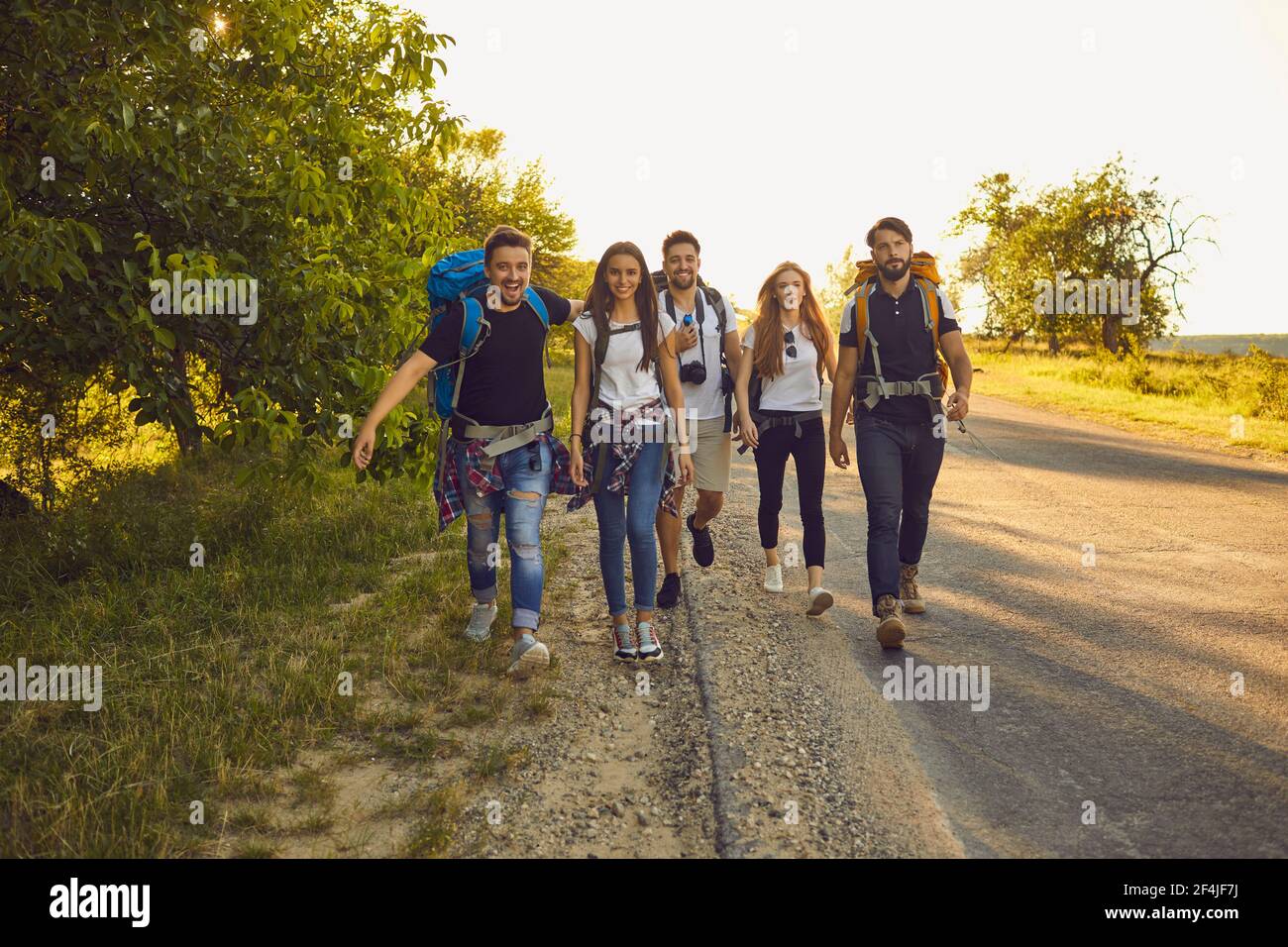 Gruppe von glücklichen jungen Freunden mit Rucksäcken entlang der Straße in der Landschaft. Wanderer auf touristischen Ausflug im Sommer Stockfoto