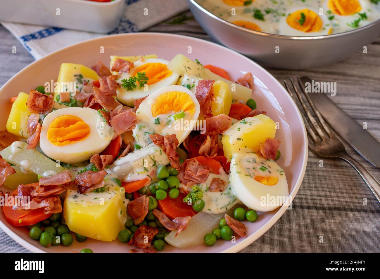 Köstliche Eierspeise mit gekochten halbierten Eiern, gebratenem Schinken, Gemüse, Kartoffeln in einer leckeren Kräutersauce serviert auf einem Teller auf dem Küchentisch Stockfoto