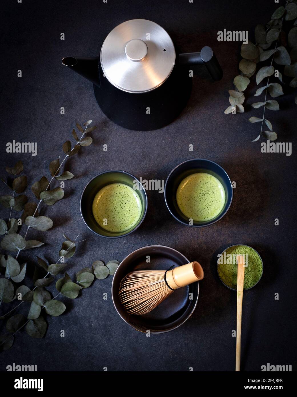 Zwei identische schwarze Teetassen auf Ton, umgeben von Eukalyptuszweigen, eine Schale macha Tee mit Bambuslöffel und Bambusbeesen auf schwarzem Hintergrund Stockfoto