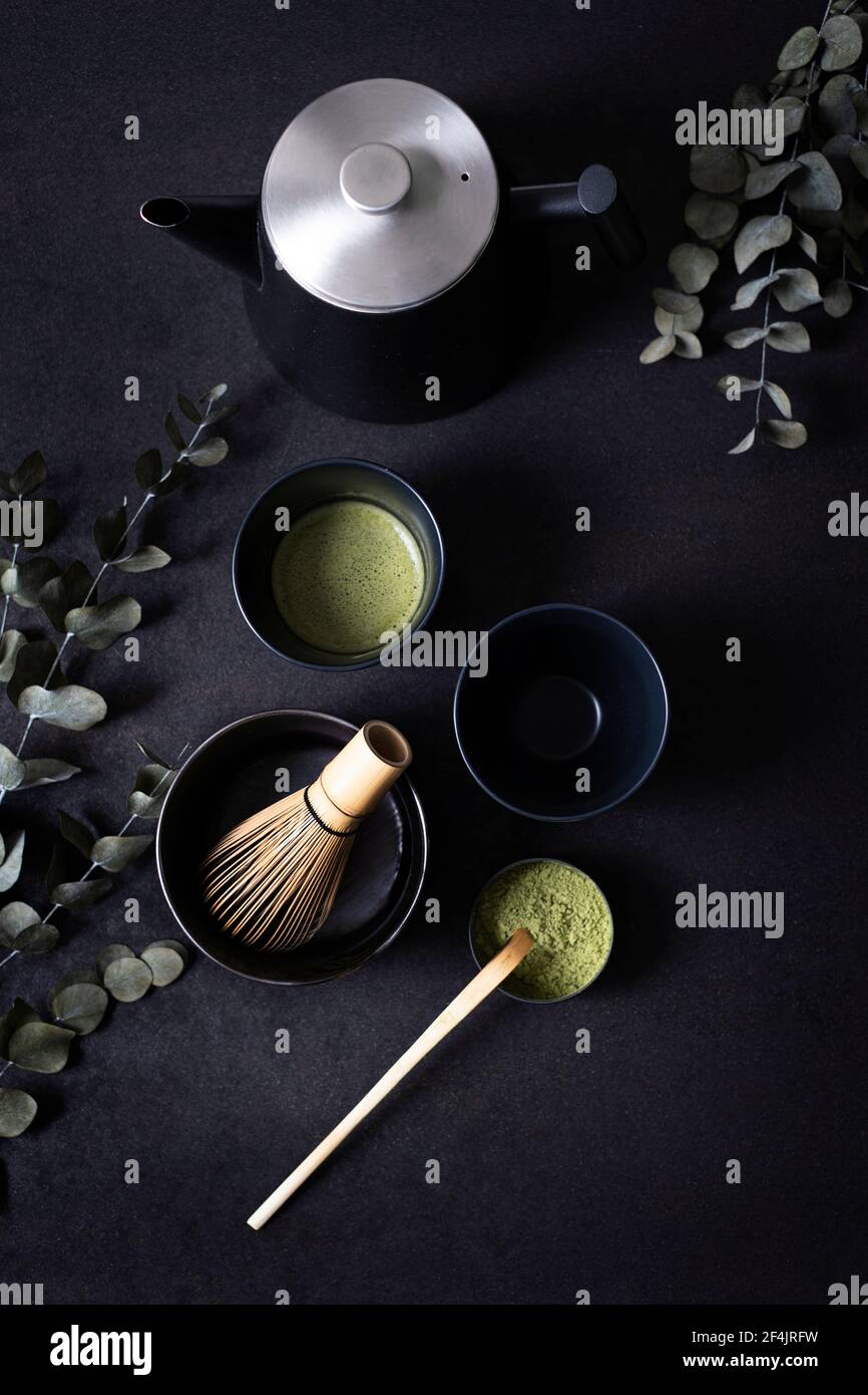 Zwei identische schwarze Teetassen auf Ton, umgeben von Eukalyptuszweigen, eine Schale macha Tee mit Bambuslöffel und Bambusbeesen auf schwarzem Hintergrund Stockfoto