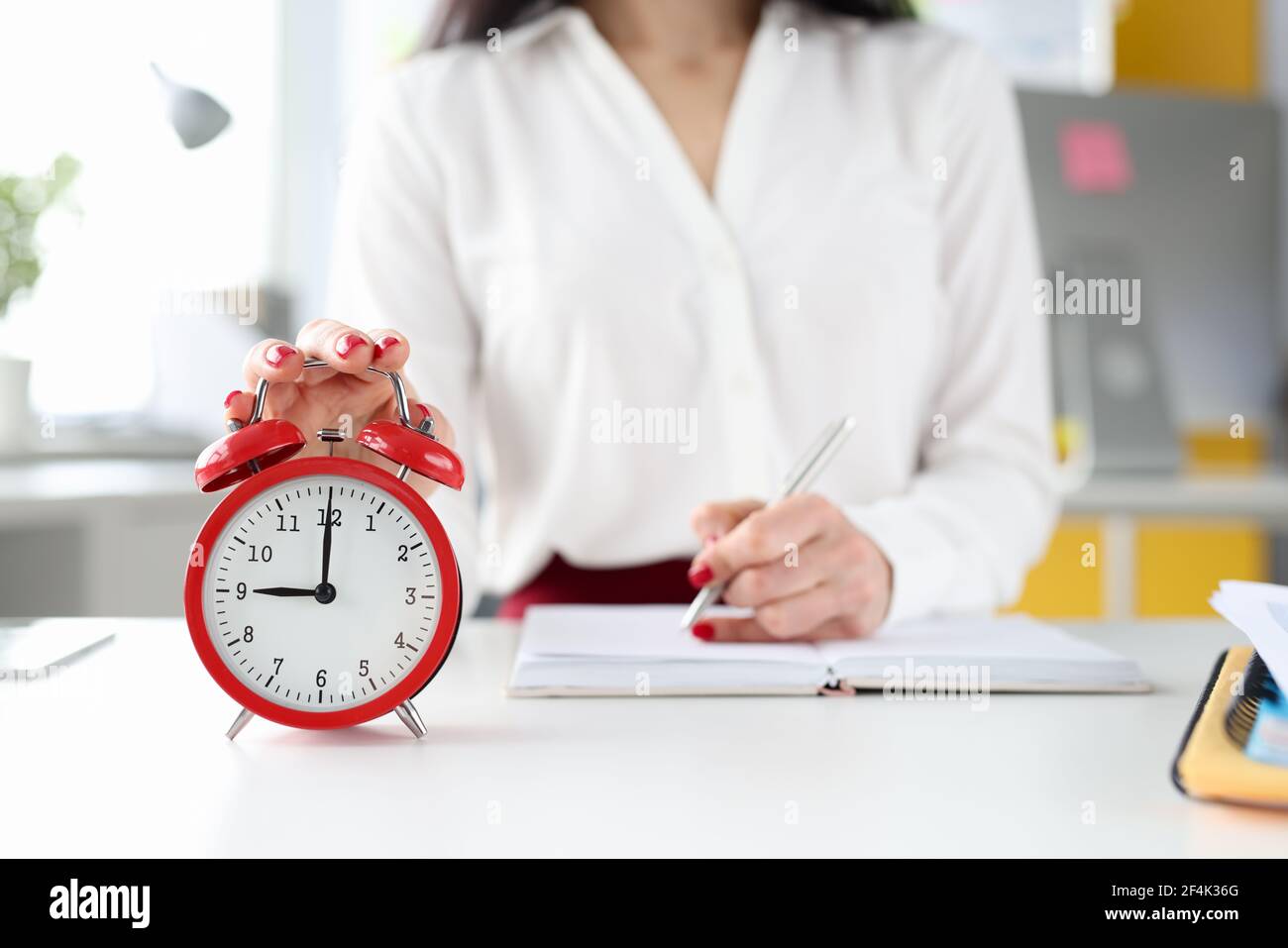 Geschäftsfrau an ihrem Schreibtisch hält ihre Hand auf rotem Alarm Uhr und nimmt Einträge in Tagebuch Stockfoto