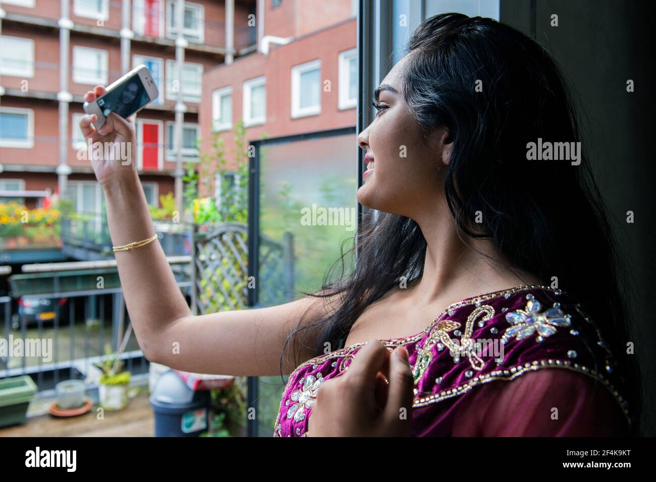 Rotterdam, Niederlande. Junge, schöne und attraktive Hindustan-Studentin in traditioneller Garderobe, die Selfies mit dem Smartphone macht, während sie in ihrer Wohnungstür steht. Stockfoto
