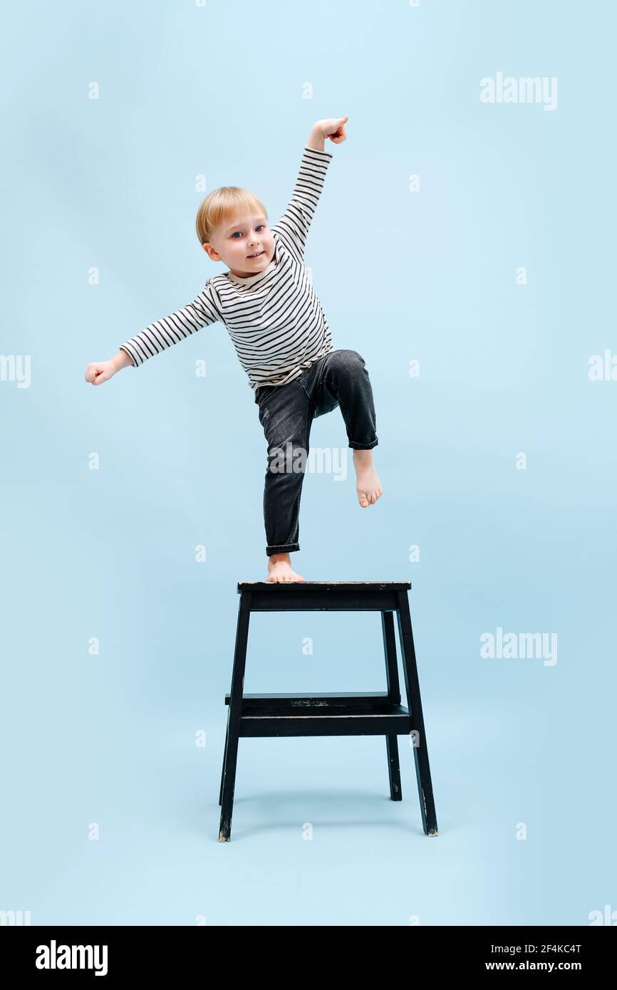 Agile barfuß blonde Junge balanciert auf einem Fuß auf einem Tritthocker, kippen zur Seite. Er trägt gestreiftes Langarm-Shirt. Studioaufnahme. Über blu Stockfoto