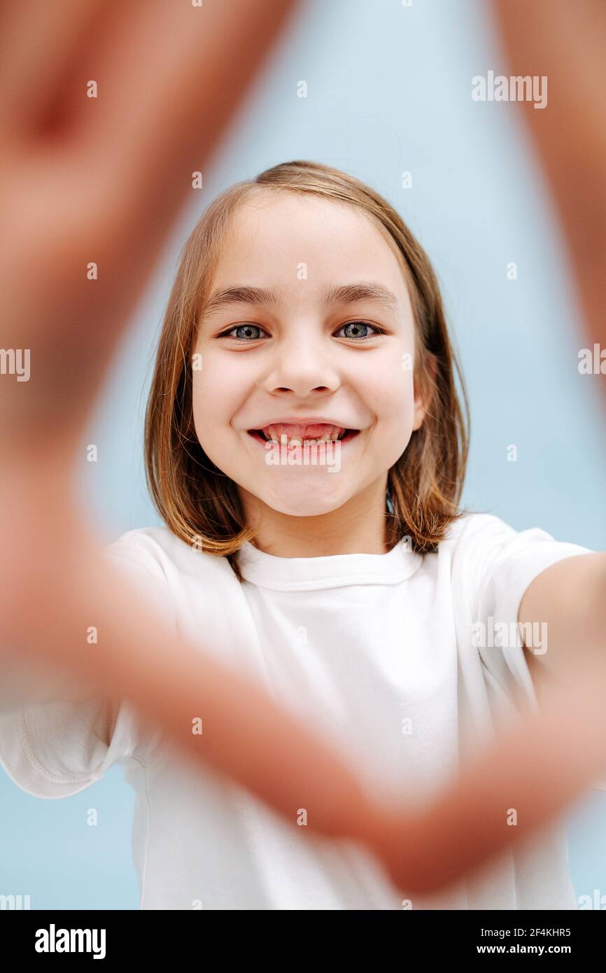 Porträt eines lächelnden 9-jährigen Mädchen durch einen Fingerrahmen macht sie. Auf blauem Hintergrund. Studioaufnahme. Sie vermisst einen Zahn. Stockfoto