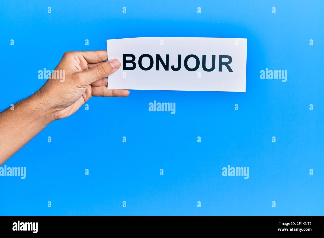 Hand von hispanischen Mann hält französisch Gruß bonjour Wort Papier über isolierten blauen Hintergrund. Stockfoto