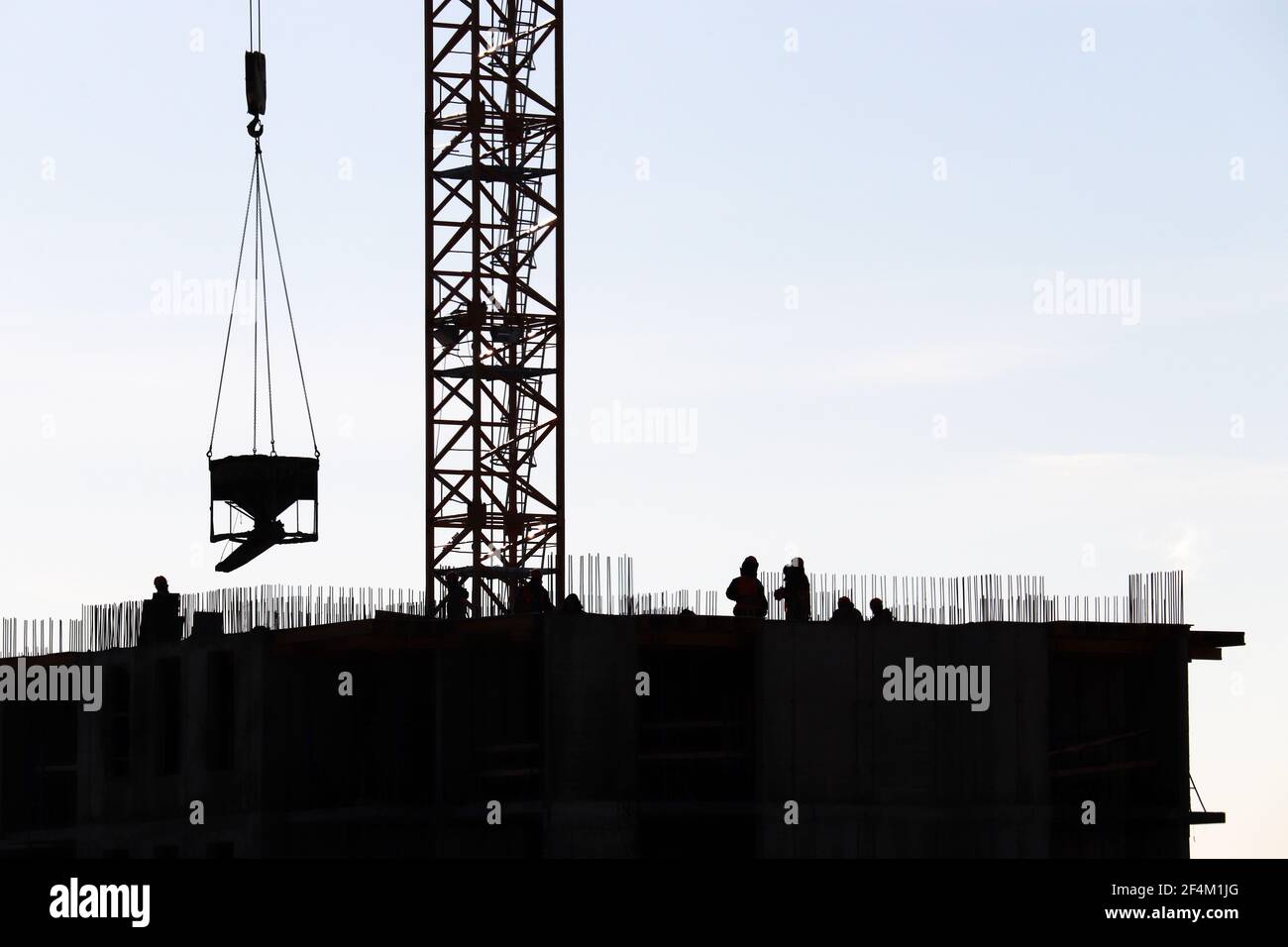 Silhouetten von Arbeitern und Turmdrehkran mit Ladung auf der Baustelle gegen den Himmel. Wohnungsbau, Bauherren arbeiten auf Gerüsten Stockfoto