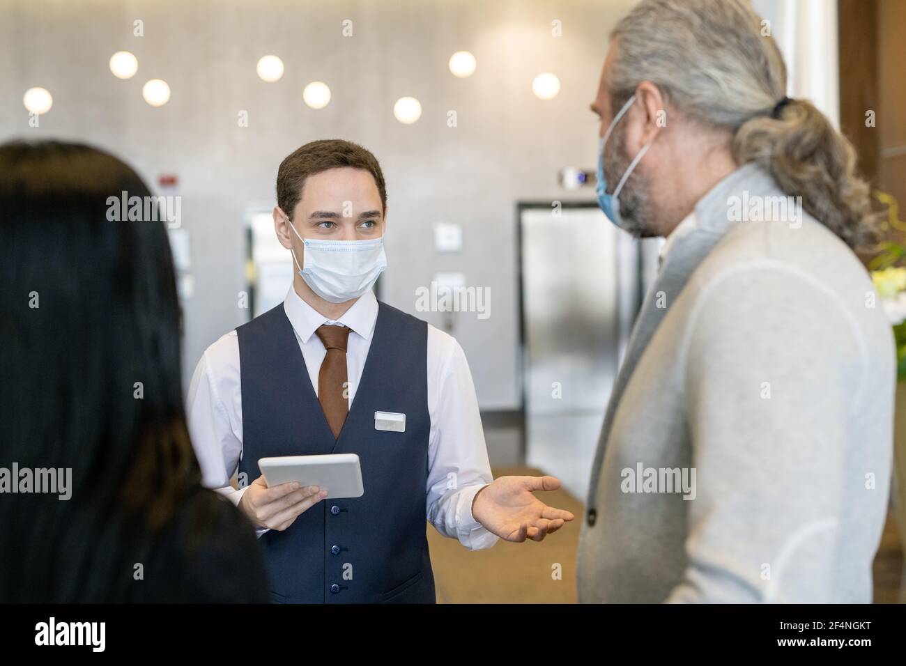 Junge männliche Rezeptionistin in Schutzmaske und formalwear mit Touchpad Im Gespräch mit einem reifen Geschäftsmann in der Lounge Stockfoto