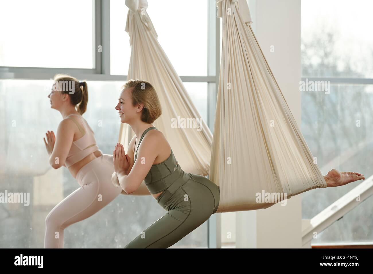 Seitenansicht von glücklichen jungen Sportlerinnen in Trainingsanzügen mit Fly Yoga-Ausrüstung während der Übung mit ihren Händen zusammen gestellt Brust Stockfoto