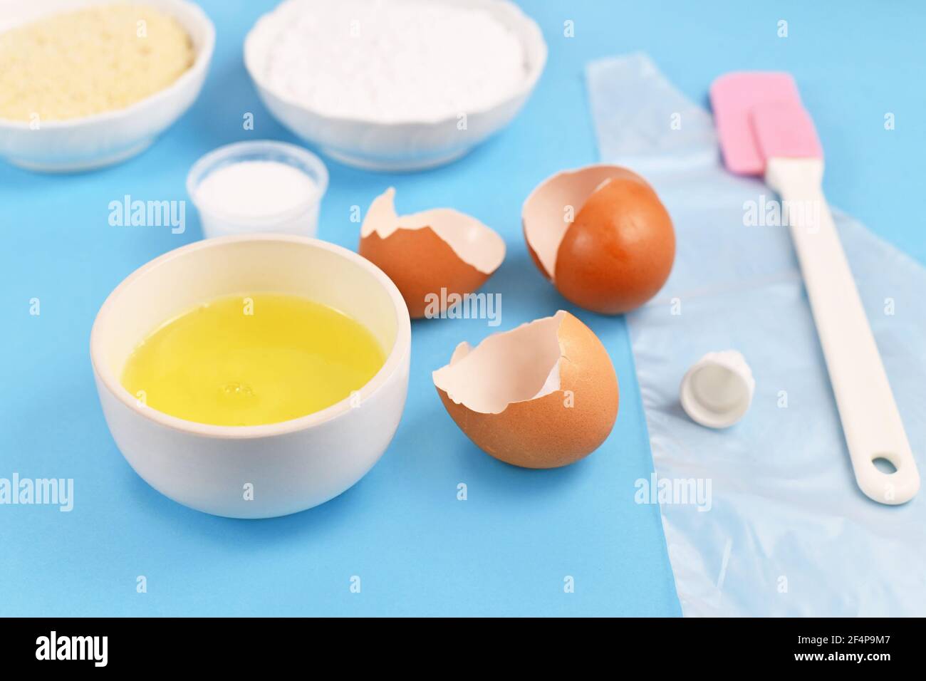 Zutaten für die Herstellung von hausgemachten französischen Macarons mit Schüssel mit Ei Weiß neben zerbrochenen Eierschalen auf blauem Hintergrund Stockfoto