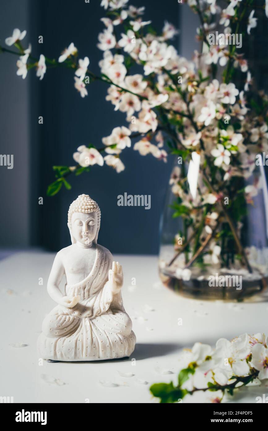 Dekorative weiße Buddha Statuette mit blutigen Ästen in der Vase auf dunklem Hintergrund. Meditations- und Entspannungsritual. Buddha Geburtstag Stockfoto