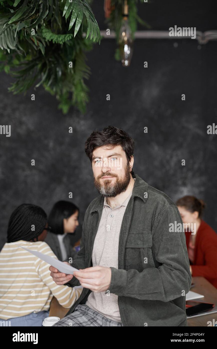 Vertikale Taille bis Porträt von zeitgenössischen bärtigen Geschäftsmann Blick auf Kamera mit verschiedenen Gruppen von Menschen treffen im Hintergrund Stockfoto