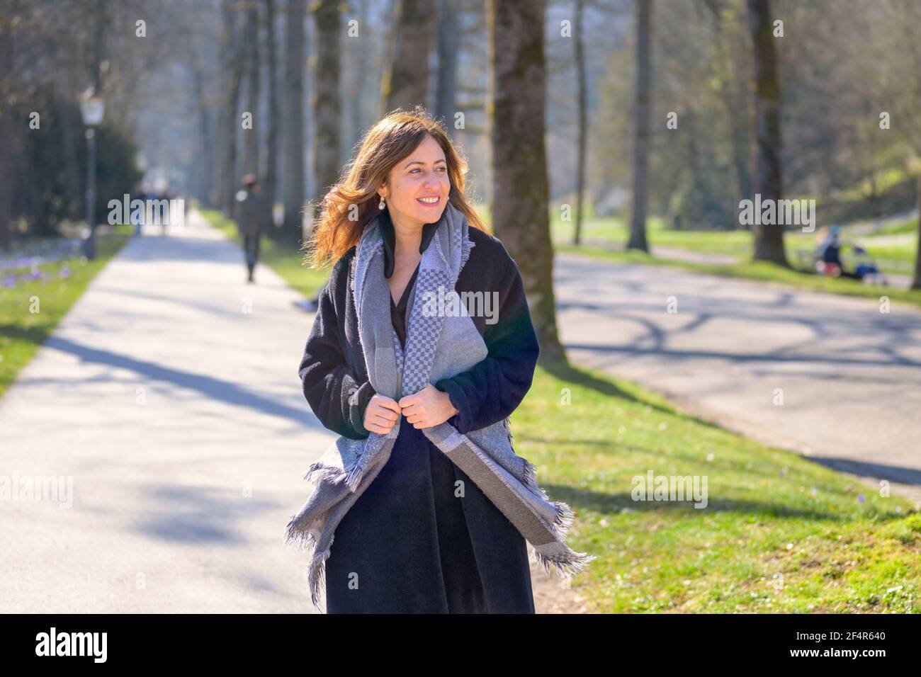 Attraktive Frau mittleren Alters lächelt zu sich selbst, als sie durch schlendert Ein Frühlingspark in einem warmen Mantel und Schal nähert sich Die Kamera genießt die Sonnen Stockfoto