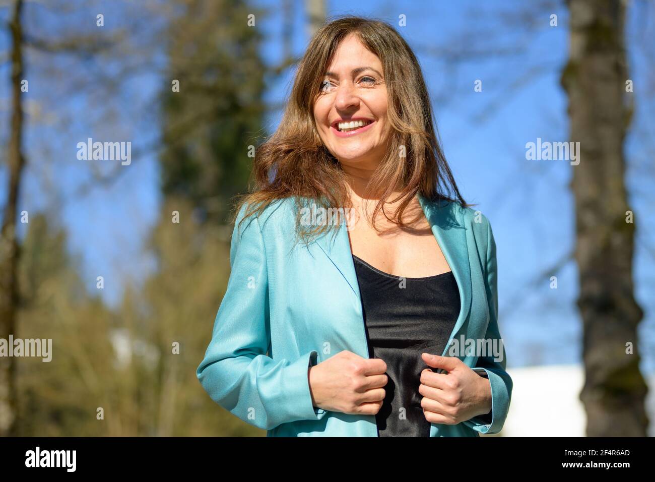 Glückliche Frau mittleren Alters in einem stilvollen blauen Jacke mit einem Fröhliches freundliches Lächeln in einer Nahaufnahme aus dem unteren Winkel Im Frühling im Freien in einem Park Stockfoto