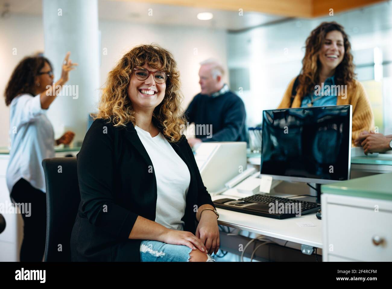 Porträt einer lächelnden Frau, die mit Menschen im Hintergrund an ihrem Schreibtisch sitzt. Gemeindeamt Rezeptionist Blick auf Kamera und lächelnd. Stockfoto