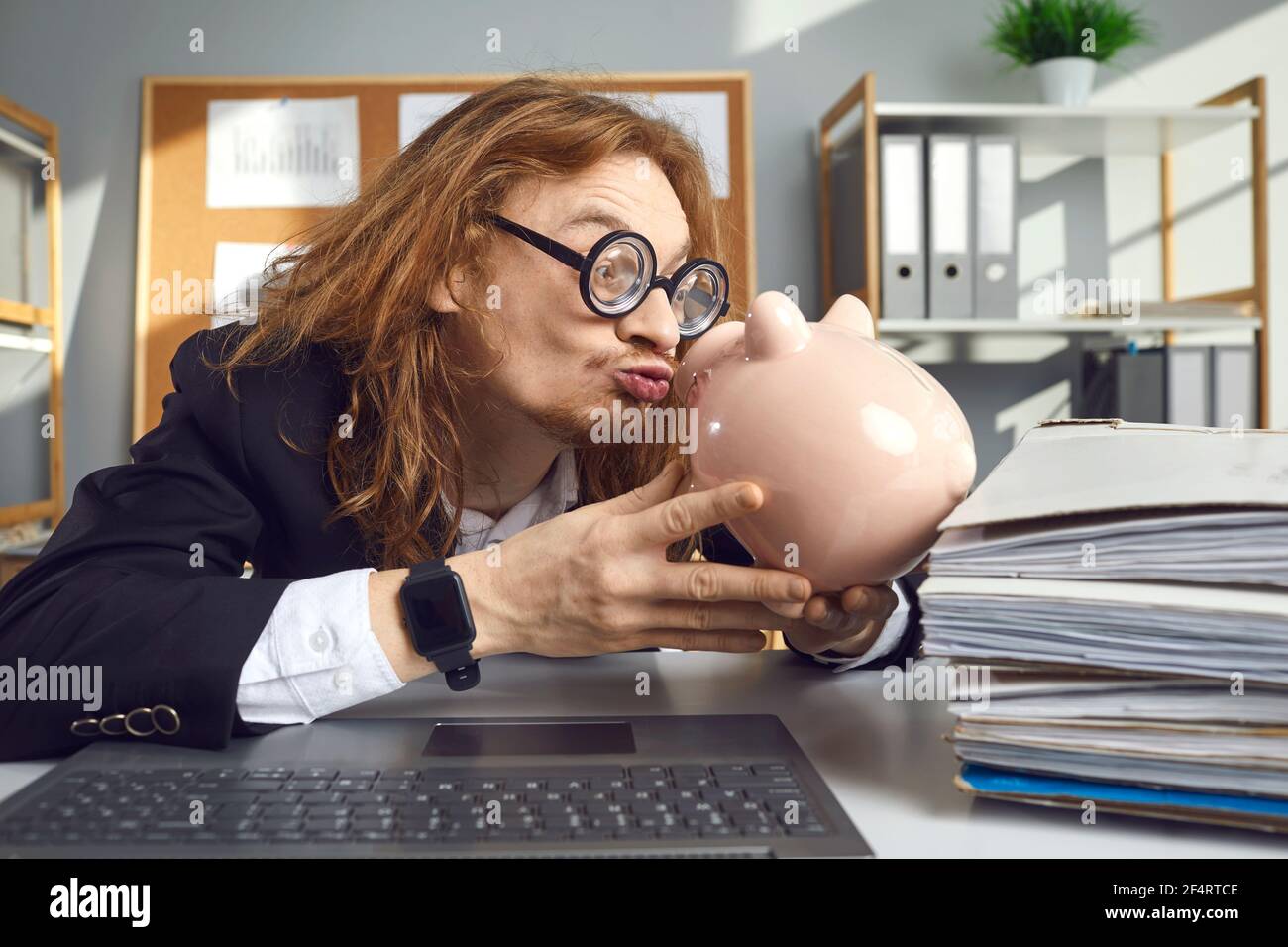 Komisch aussehender Mann in lustigen Gläsern, der am Schreibtisch sitzt Und küssen Sparschwein Stockfoto