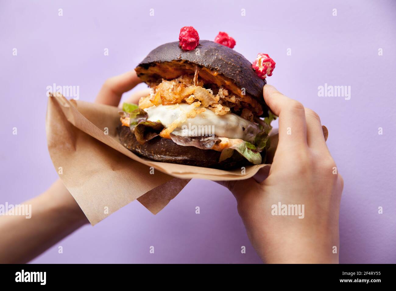 Corona (antiCOVID) Cheeseburger mit hausgemachten schwarzen Brötchen, Salat, gebratene Zwiebel und Käse in weiblichen Händen auf violettem Hintergrund. Restaurant, Burger Bar Stockfoto