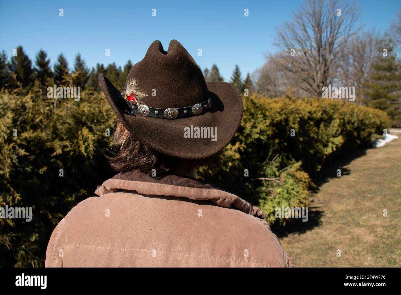 Eine Frau mit kurzen braunen Haaren trägt einen braunen Cattleman Cowboy Hut mit einer Feder und dekorativen Gürtel befestigt. Blick auf Bäume. Stockfoto