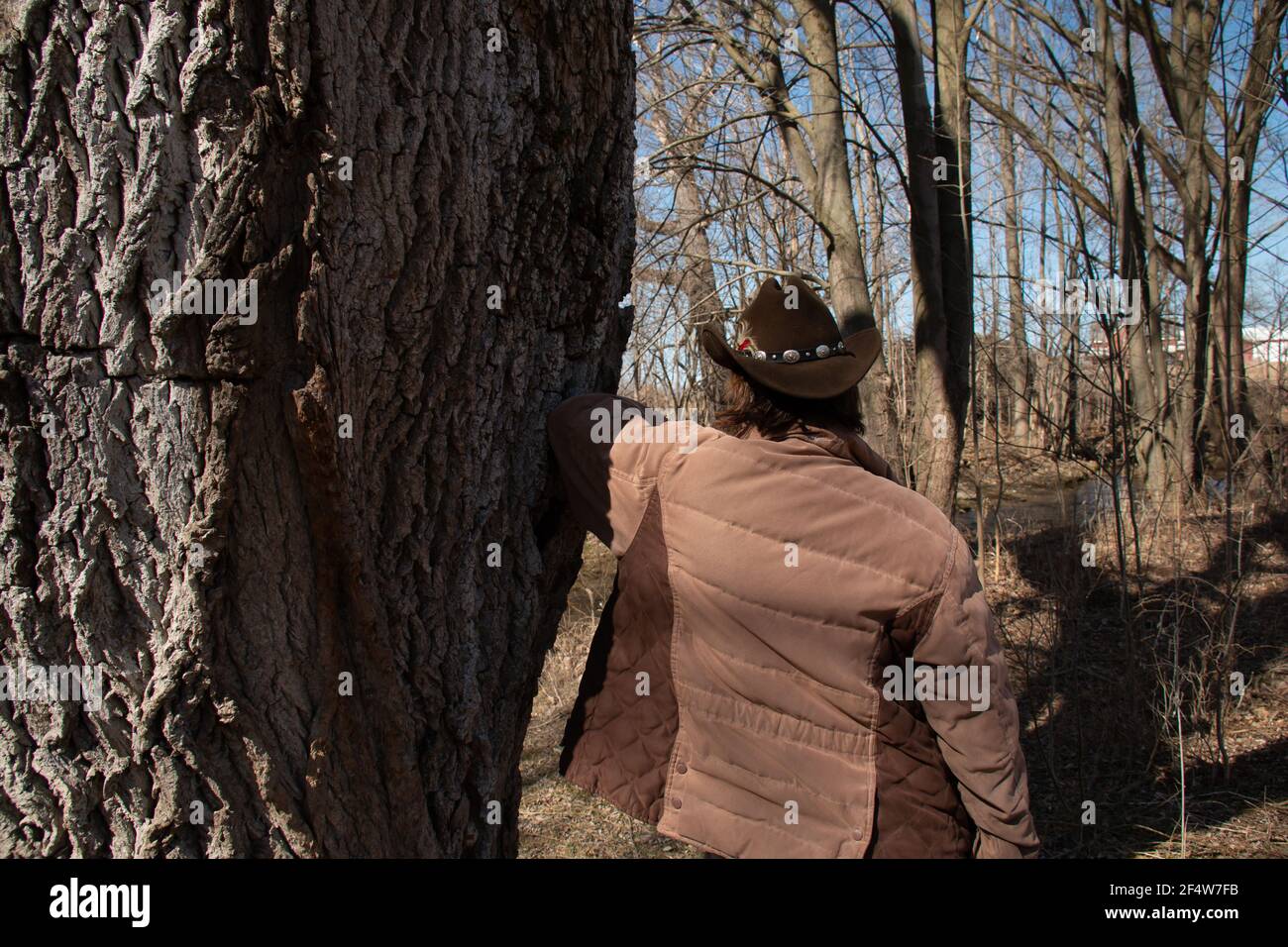 Eine Frau mit kurzen braunen Haaren trägt einen braunen Cattleman Cowboy Hut mit einer Feder und dekorativen Gürtel befestigt. Lehnt sich an einen Baum Stockfoto
