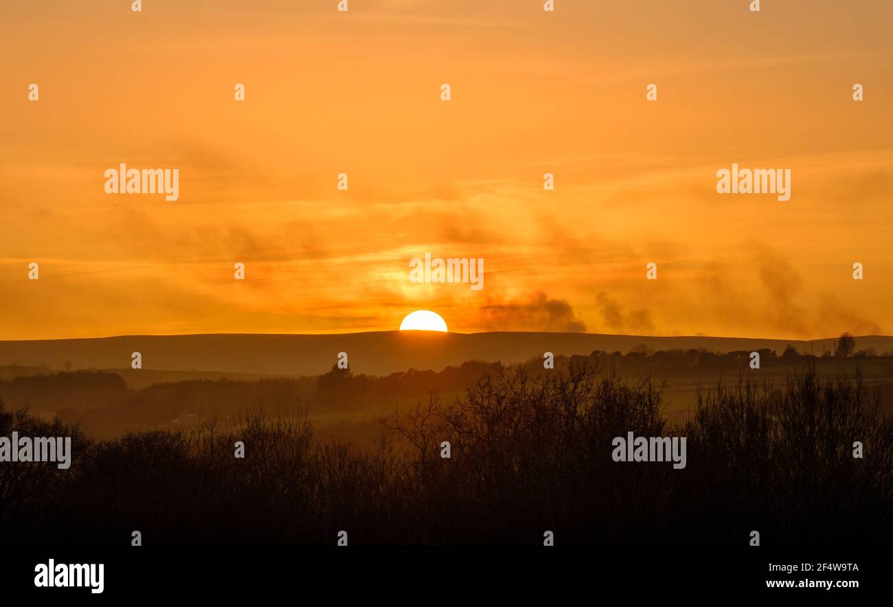 Nebliger orangefarbener Sonnenuntergang über den North Durham Moors am östlichen Rand der Pennines. Rauchen von brennender Heide, die über die untergehende Sonne driftet Stockfoto